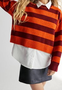 Gestreepte gebreide trui in oranje en bordeaux, met een cropped pasvorm en ribgebreide zoom. Gecombineerd met een witte blouse met kraag en een zwarte leren rok.
