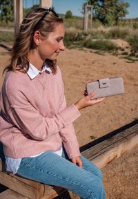 Žena v růžovém svetru sedí venku na dřevěné lavici, drží v pravé ruce světle šedou peněženku a dívá se na ni.