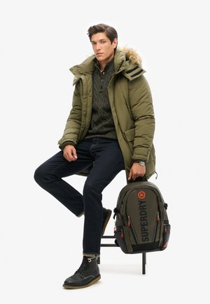 Jeune homme assis sur un tabouret portant une parka verte, un pull sombre, un jean et des bottes noires, tenant à ses côtés un sac à dos Superdry vert assorti.