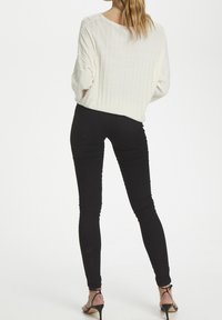 Weißer, gerippter Strickpullover, schwarze Skinny-Jeans und schwarze Stiletto-Heels. Bemerkenswerte Details: lockere Passform des Pullovers und knöchellange Jeans.