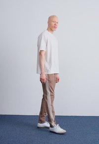 Lichtgrijs shirt met korte mouwen, beige joggingsbroek met witte zijkanten, en witte sneakers gedragen door een staand persoon op een blauwe vloerbedekking.