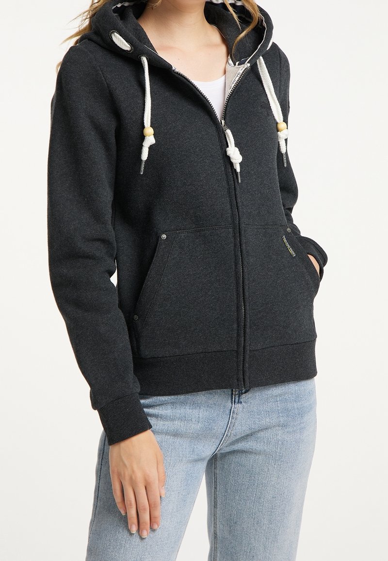 Femme portant un sweat à capuche zippé gris foncé avec des cordons blancs et un jean bleu clair, debout avec une main dans la poche.