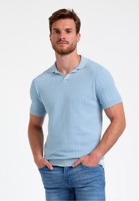 Gaiši zils, īsu piedurkņu polo krekls ar ribotu tekstūru un pogājamu apkakli. Ikdienas piegriezums ar taisnu apakšmalu. Nēsāts ar džinsu biksēm.