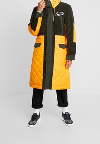 Vadderad lång jacka med orange- och olivgröna sektioner, texturerad fleece, huva, dragkedjestängning och sidofickor. Logotyp på bröstet.