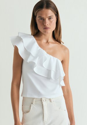 Mujer con top blanco de un hombro con detalle de volante y pantalones de color claro, mirando hacia adelante con expresión neutra.