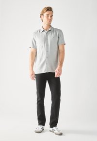 Camisa de manga corta gris claro con cierre de botones y un logo sutil, combinada con jeans negros y zapatillas blancas con rayas negras.