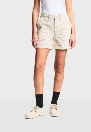 Model nosi bež kratke hlače s manšetama i prednjim džepovima s zakrpama, crne učvršćene čarape i bež cipele s vezicama, stoji ispred jednostavne pozadine.