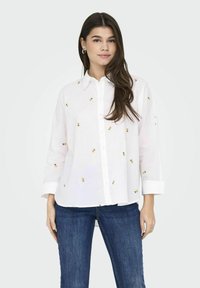 Camicia bianca con ricamo di piccoli ananas gialli, maniche lunghe e vestibilità rilassata, abbinata a jeans in denim blu.