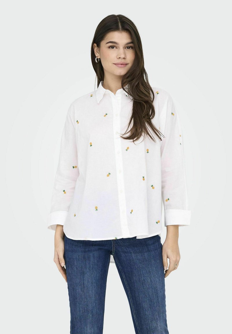 Camicia bianca con ricamo di piccoli ananas gialli, maniche lunghe e vestibilità rilassata, abbinata a jeans in denim blu.
