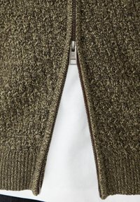 Hnedý a béžový pletený sveter na zips čiastočne rozopnutý cez bielu košeľu, ktorý ukazuje textúru a detaily zipsu.