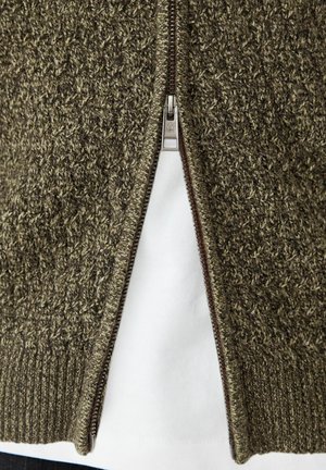 Hnědý a béžový pletený svetr na zip částečně rozepnutý přes bílou košili, zobrazuje texturu a detaily zipu.