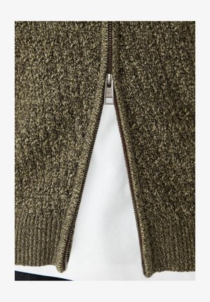 Hnědý a béžový pletený svetr na zip částečně rozepnutý přes bílou košili, zobrazuje texturu a detaily zipu.