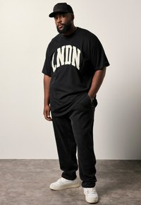 Sort grafisk t-shirt med "LNDN" i store cremefarvede bogstaver, parret med mørke jeans og hvide sneakers. Modellen bærer en sort kasket.