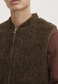 Brun fleece vest med lynlås foran og høj krave. Strukturert stof suppleret med en brun langærmet skjorte under.