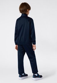 Granatowy dres z gładkiego, błyszczącego materiału, z elastycznymi mankietami i pasem, połączony z ciemnymi sneakersami z niebieskimi akcentami.