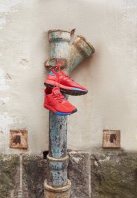 Czerwone buty sportowe z niebieskimi akcentami, z przewiewną cholewką i teksturowaną podeszwą, wiszące na zniszczonej, rusty metalowej rurze na tle ściany.