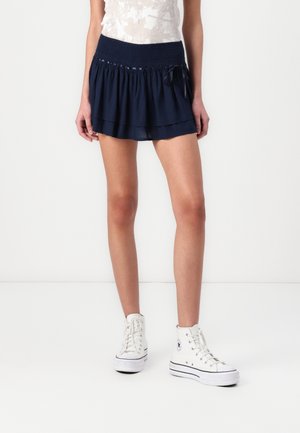Kvinde iført en mørkeblå lagdelt miniskørt med en smock-bånd i taljen og hvide høje sneakers, stående mod en hvid baggrund.