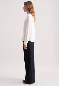 Donna con capelli castano chiaro e mossi in profilo, indossa una blouse bianca a maniche lunghe, pantaloni larghi scuri e scarpe nere a punta.