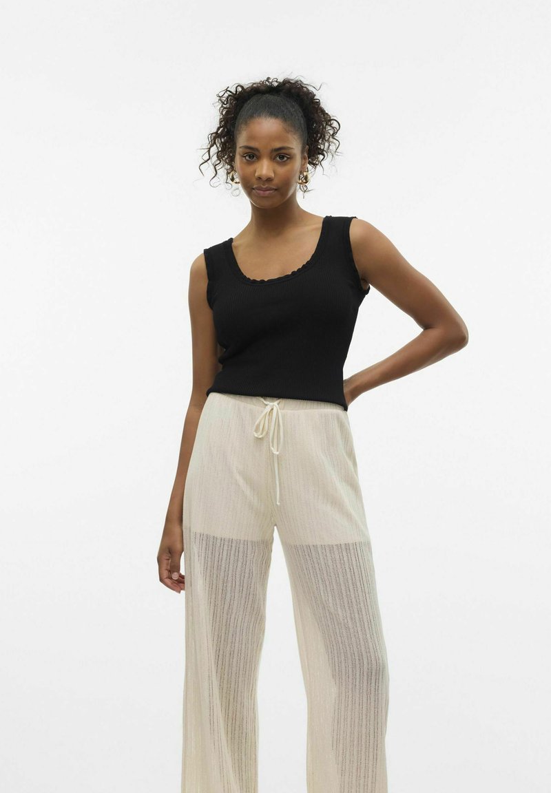 Top de tirantes negro con textura acanalada y escote con volantes, combinado con pantalones amplios claros, transparentes y plisados que cuentan con una cintura con cordón.