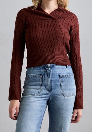 Frau trägt einen braunen, kurz geschnittenen Pullover mit Zopfmuster und geripptem Saum sowie hellblaue, hoch taillierte Jeans mit großen Vordertaschen.