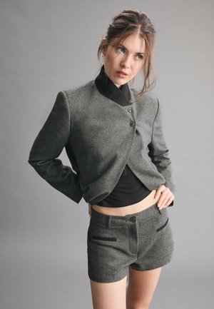 Jeune femme portant un blazer gris et un short assorti avec un haut noir, posant les mains sur les hanches devant un fond gris uni.
