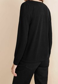 Černý dlouhý rukáv top vyrobený z měkké látky, s kulatým výstřihem a přiléhavými rukávy, nošený s černými kalhotami, z pohledu zezadu.