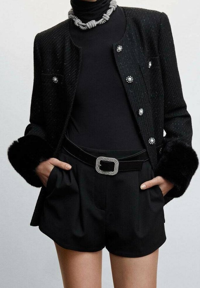Veste noire texturée avec boutons en perles et poignets en fourrure, portée sur un col roulé noir et un short taille haute avec une boucle de ceinture ornée de bijoux.