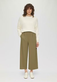 Crème gebreide trui met geribbelde textuur, gecombineerd met olijfgroene wijde broek. Model draagt witte sneakers, met handen in de zakken, neutrale achtergrond.