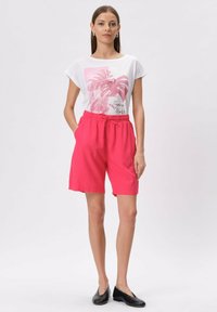 Biała koszulka z różowym tropikalnym nadrukiem, w połączeniu z jasnoróżowymi szortami z elastycznym paskiem. Modelka ma na sobie czarne baleriny.
