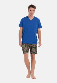 Blå bomulds t-shirt med en lomme, parret med farverige shorts med blomstermønster i nuancer af pink, grøn og gul. Barfodet.