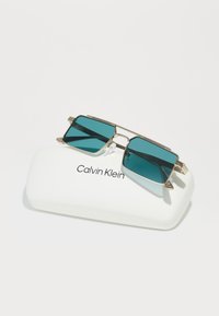 Occhiali da sole Calvin Klein con lenti teal, montatura rettangolare in metallo e naselli trasparenti, appoggiati su un astuccio bianco marchiato.