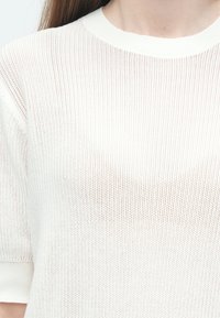 Haut blanc tricoté avec un col rond, présentant une texture ajourée ample et des manches courtes. Tissu doux et léger.