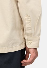 Beige skjorte med knaplukning og struktureret overflade, med en manchet der lukkes med knap og stikninger på ærmerne.