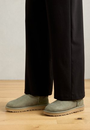 Botas de tornozelo em camurça verde com bico redondo e sola de borracha, combinadas com calças pretas largas, sobre um chão de madeira.
