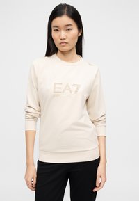 Μπεζ μακρυμάνικη μπλούζα με στρογγυλή λαιμόκοψη, διακοσμημένη με ένα χρυσό λογότυπο στο μπροστινό μέρος, με ελαστικές μανσέτες και τελείωμα. Ομαλή υφή.