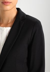 Černý blejzr se strukturovaným límcem, hladkým materiálem a přiléhavým střihem, nošený přes bílé haleně s knoflíkovým zapínáním.