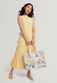 Abito giallo senza maniche con gonna plissettata. Borsa a mano con motivo floreale in colori pastello e manici crema, abbinata a ballerine oro metallizzato.