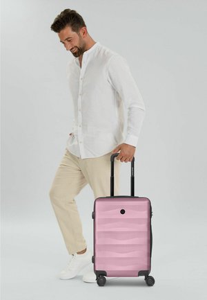 Homme en chemise blanche et pantalon beige tirant une valise rigide rose à poignée extensible, debout sur fond gris.