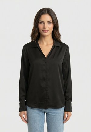 Donna con capelli castani fino alle spalle che indossa una camicia nera a maniche lunghe con bottoni e jeans azzurri, in piedi davanti a uno sfondo uniforme.