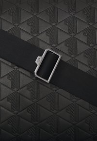 Svart teksturert veske med geometrisk mønster, accentuert med svart stropp og rektangulær metallspenne.