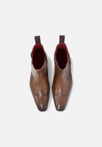 Botas de tobillo de cuero marrón con un acabado pulido, que presentan detalles de brogue, punta afilada y forro interior rojo. Etiquetas en el lazo para facilitar el calzado.