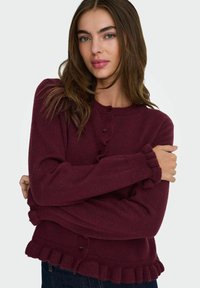 Gilet en maille bordeaux avec un col rond, fermeture à boutons et ourlet volanté. Texture douce et manches longues avec poignets volantés.