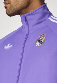 Μωβ αθλητικό σακάκι με ψηλό γιακά και κλείσιμο με φερμουάρ. Φέρει κεντημένο λογότυπο της Ρεάλ Μαδρίτης και ανακλαστικό τριφύλλι της Adidas στο αριστερό στήθος.