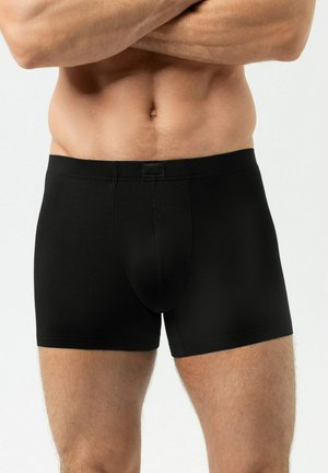 Fit mannelijk torso met gekruiste armen, gekleed in zwarte strakke boxer shorts tegen een eenvoudige lichte achtergrond.