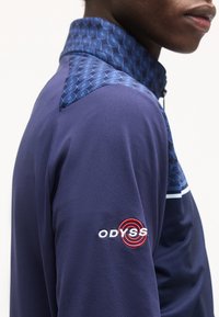 Κοντινό πλάνο ενός ατόμου που φοράει σκούρο μπλε μπουφάν με γιακά με σχέδιο και το λογότυπο "ODYSSEY" κεντημένο στο μανίκι σε λευκό και κόκκινο.