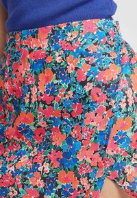 Jupe florale à imprimé multicolore présentant des fleurs rouges, bleues et oranges sur un fond noir. Tissu léger avec une fente sur le côté.