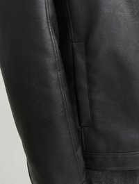 Detalle de una chaqueta de cuero negra que presenta una textura suave, un ajuste entallado y un bolsillo lateral. La costura es visible a lo largo de los bordes.