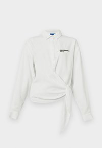 Camisa blanca de manga larga con un diseño envolvente, tapeta de botones, cuello puntiagudo y logotipo negro bordado en el pecho. Textura de algodón suave.