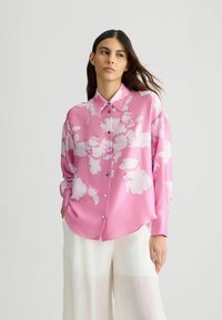 Lange mouwen roze blouse met een wit bloemenpatroon, met een kraag met knopen en knopen aan de voorkant, gemaakt van gladde stof, gecombineerd met lichte broeken.