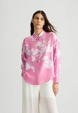 Blusa rosa a maniche lunghe con motivo floreale bianco, colletto a camicia, con bottoni davanti, realizzata in un tessuto liscio, abbinata a pantaloni chiari.